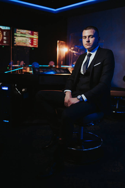 A vertical shot of a young caucasian man in a black suit sitting on a chair in a casino - Photo, Image