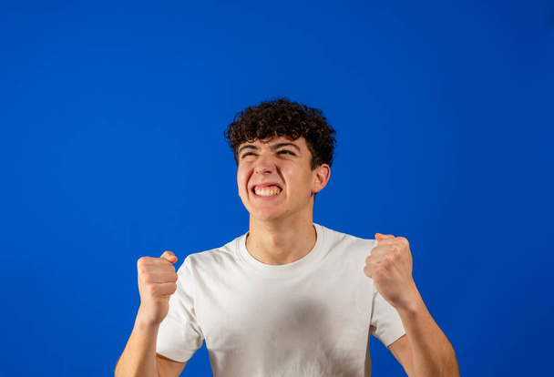Attraktiver junger Mann mit lockigem Haar, der wütend die Fäuste ballt, isoliert auf blauem Studiohintergrund. Konzept der Rebellion - Foto, Bild