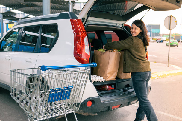 усміхнена жінка поклала сумки з продуктами в багажник автомобіля після продовольчого магазину копіювання простору
 - Фото, зображення
