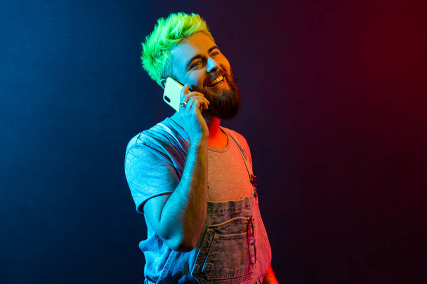 Portrait of young adult hipster man wearing denim overalls and t shirt talking phone, hears happy news, smiles toothily, expressing optimism. Colorful neon light, indoor studio shot. - Foto, Imagen