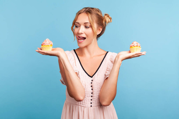 Young adult adorable blonde female with two hair buns wearing dress holding two cakes in hands, showing tongue, wants to lick and eat dessert. Indoor studio shot isolated on blue background. - Foto, Bild