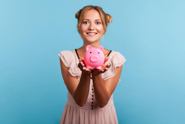 Attractive smiling young adult woman with pleasant appearance and blonde hair, showing piggy bank to camera, happy with money savings. Indoor studio shot isolated on blue background. - Foto, afbeelding