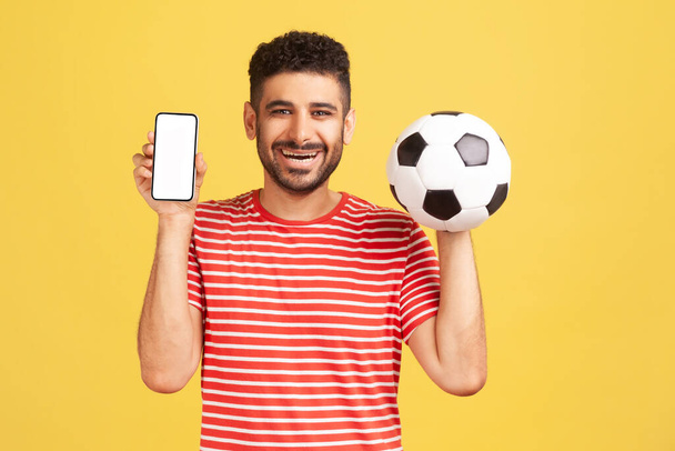 Smiling positive man with beard in striped t-shirt holding and showing white display smartphone and football ball, betting on soccer. Indoor studio shot isolated on yellow background - Foto, afbeelding