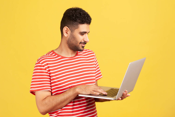 Bearded man freelancer in red striped t-shirt standing holding laptop, typing on keyboard, doing his job, teleworking on wireless gadget. Indoor studio shot isolated on yellow background - Fotoğraf, Görsel