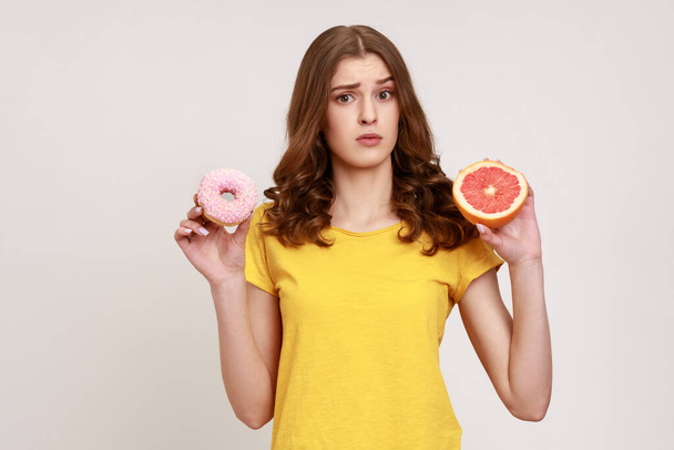Sad teenager girl holding healthy fruits and high-calorie dessert, confused wavy-haired female in T-shirt choosing between grapefruit and donut. Indoor studio shot isolated on gray background. - Fotó, kép
