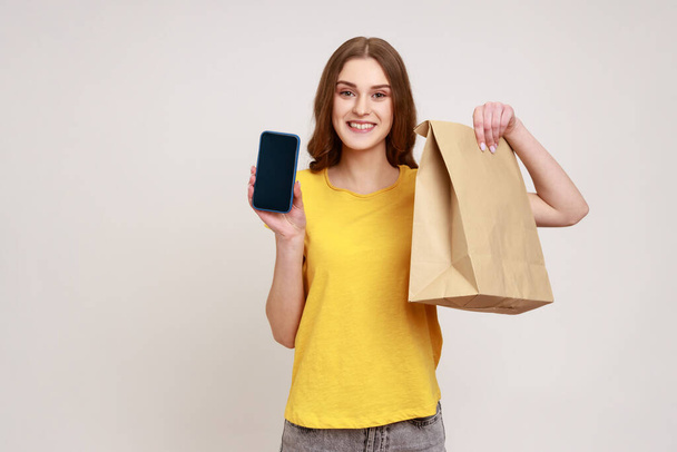 Smiling positive teenager girl in casual yellow T-shirt holding blank display smartphone and eco paper bag, online shopping, fast delivery, marketplace. Indoor studio shot isolated on gray background. - Foto, imagen