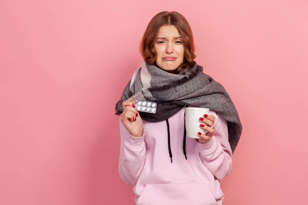 Portrait of ill curly haired teenage girl in hoodie and scarf making miserable face and holding tea cup and pills, treatment. Indoor studio shot isolated on pink background - Photo, image
