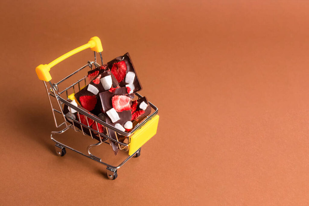 Trendy still life composition with toy shopping cart and natural organic chocolate with sublimated strawberries, on brown paper background with copy space. Composition for sugar-free chocolate brand - Photo, Image