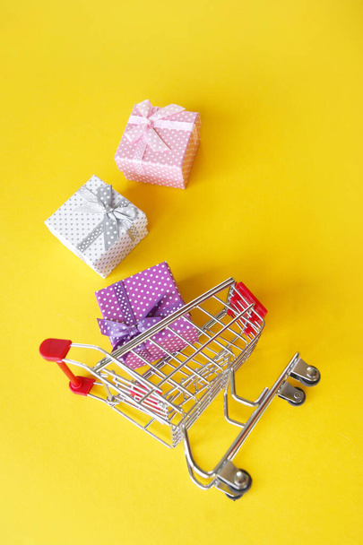 Mini ostoskori pieniä lahjoja keltaisella taustalla. Lahjojen ostamisen käsite myymälöissä ja Internetissä. - Valokuva, kuva