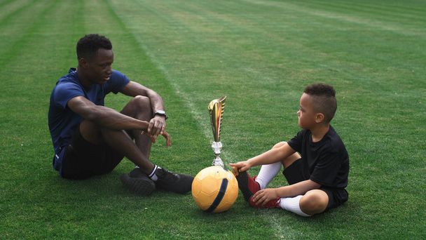 Diverso padre e hijo hablando cerca de la copa de oro en el campo de fútbol - Foto, Imagen