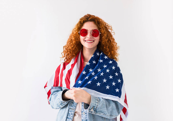 Mujer joven bonita feliz con gafas sostienen una bandera, aislado sobre fondo blanco. La fiesta patriótica y la celebración el día de la independencia de los EEUU 4 de julio. - Foto, imagen