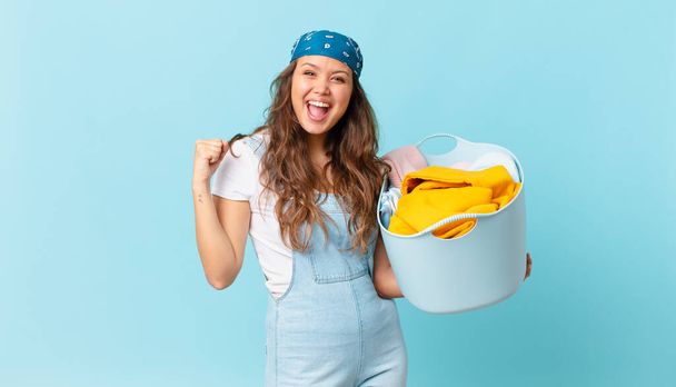 young pretty woman feeling shocked,laughing and celebrating success and holding a wash clothes basket - Foto, afbeelding
