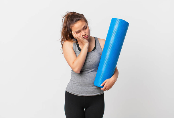 young pretty woman feeling bored, frustrated and sleepy after a tiresome and holding a yoga mat - Фото, изображение