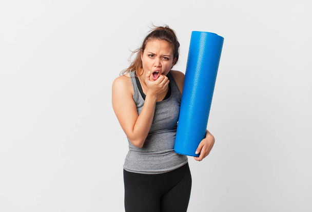 young pretty woman with mouth and eyes wide open and hand on chin and holding a yoga mat - Foto, imagen