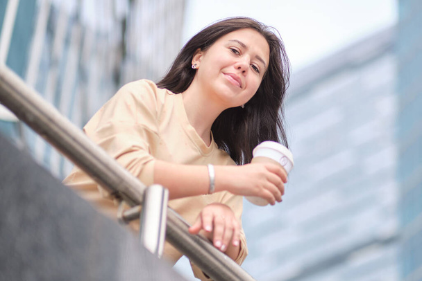 όμορφη νεαρή γυναίκα απολαμβάνει ένα φλιτζάνι καφέ, με φόντο ουρανοξύστες Χαμογελώντας hipster κορίτσι με φλιτζάνι ζεστό ρόφημα στην οδό της πόλης. - Φωτογραφία, εικόνα