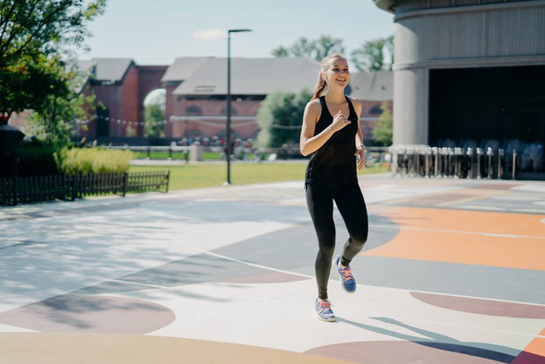 Cheerful athletic woman runs actively on stadium dressed in black sportclothes enjoys physical activities outdoors during summer day being full of energy. People sport and motivation concept - Foto, Bild