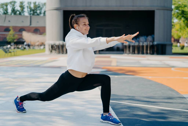 Outdoor shot of slim sporty woman involved in recreation activities stretches arms legs looks into distance wears white hoodie leggings and sportshoes stetches body before run leads healthy lifestyle - Photo, Image