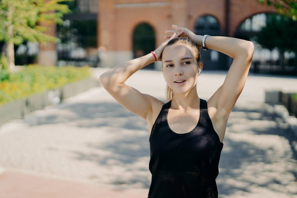 Thoughtful sporty woman concentrated into distance breathes deeply after jogging takes break during cardio training dressed in black t shirt keeps hands on head listens audio track in earphone - Foto, immagini