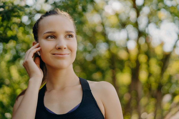 Healthy lifestyle concept. Outdoor shot of sportswoman enjoys favorite playlist in earbuds while training in park has gentle smile concentrated into distance poses over blurred nature background - Valokuva, kuva