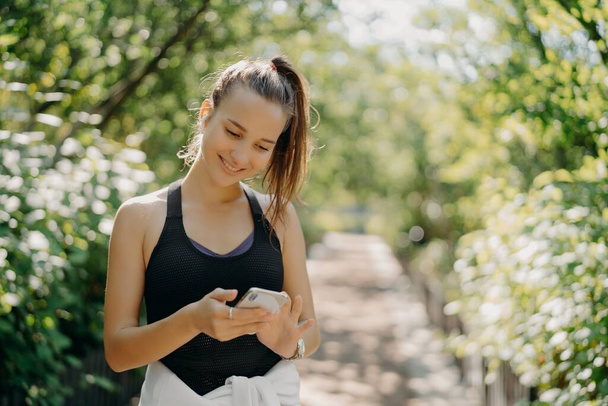 Outdoor shot of happy athletic young woman uses smartphone sport tracker application uses modern device for health and fitness being smart about training poses against blurred nature background - Photo, Image