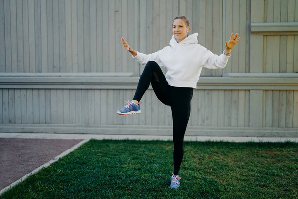 Energetische sporter heeft workout routine poses op een been demonstreert haar uithoudingsvermogen opwarmt voordat joggen gekleed in hoodie leggings en trainers traint buiten. Sport en motivatie - Foto, afbeelding