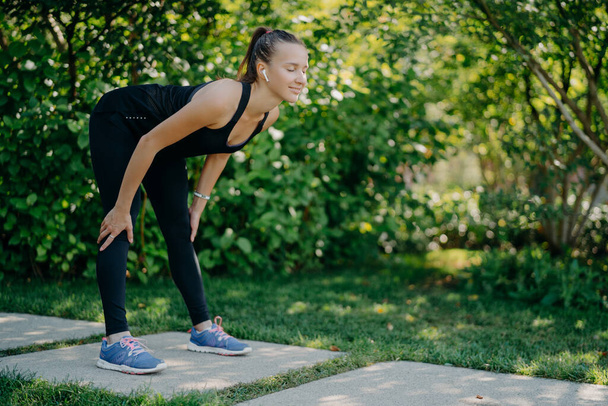 Fitness-Frau in schwarzer Sportkleidung hält die Hände auf Knien lehnt sich nach vorne Übungen im Freien vor grünem Naturhintergrund hört Musik von Lieblings-Playlist. Gewichtsverlust und Trainingskonzept - Foto, Bild