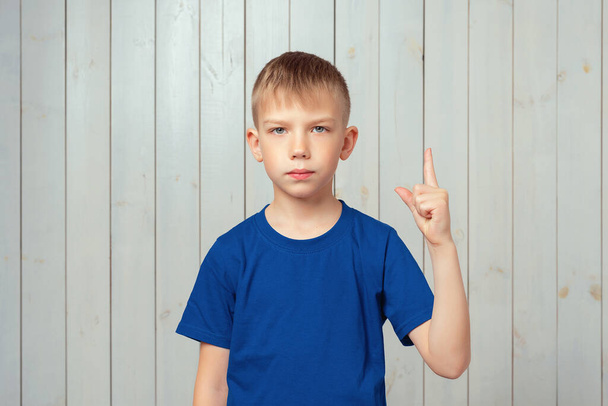 Portret van een serieuze knappe jongen in een blauw t-shirt, wijzend met de vinger omhoog en advies gevend, zegt Aandacht alsjeblieft, luister naar me. Studio shot, lichte houten achtergrond - Foto, afbeelding