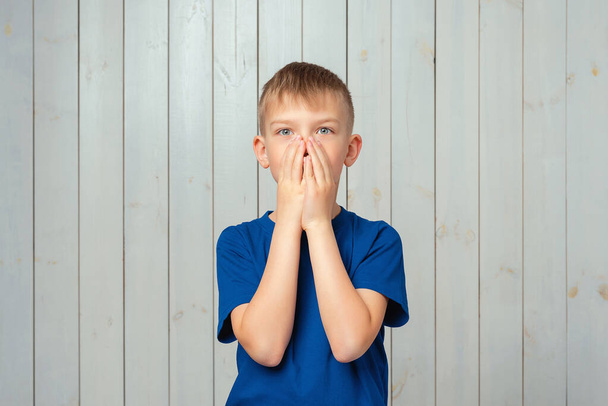 Scared preteen boy in blue t shirt hold hands near mouth trembling fear, very terrified over scary creepy sound, standing in stupor. Studio shot, light wooden background - Photo, Image