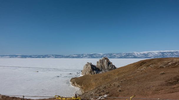 Двоголовий камінь, позбавлений рослинності, здіймається над замерзлим озером. Крихітні силуети людей видно на льоду. Гірський хребет проти блакитного неба. Байкал - Фото, зображення