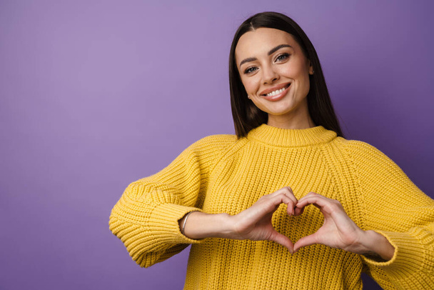 Happy young woman in sweater showing heart symbol with hands over violet wall background - Fotografie, Obrázek