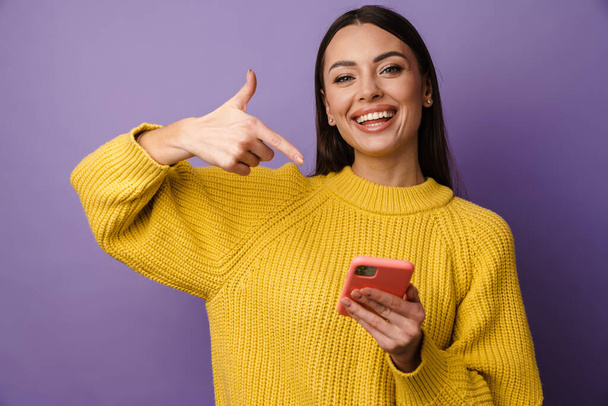 Happy young woman wearing sweater pointing finger at mobile phone over violet background - Fotoğraf, Görsel