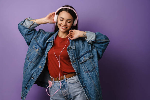 Happy young woman in denim jacket wearing headphones, dancing over violet wall background - Фото, изображение