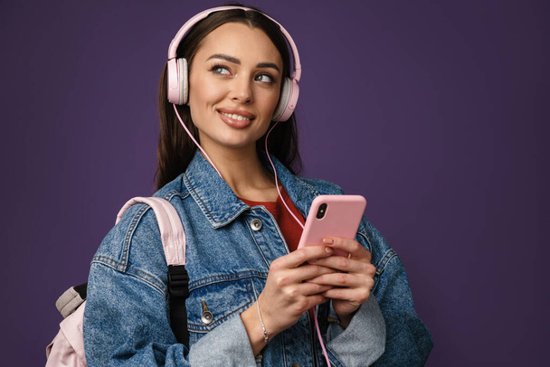 Happy young woman in denim jacket wearing headphones, holsing mobile phone over violet wall background - Фото, зображення