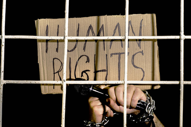 das Konzept der Menschenrechte Redefreiheit Hände mit einer eisernen Kette gefesselt halten ein Mikrofon und ein Pappschild mit der Aufschrift Menschenrechte hinter einem eisernen Gitter Gefängnis. Hochwertiges Foto - Foto, Bild