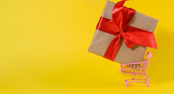 caja de regalo con un lazo de seda roja en un carro de metal en miniatura sobre un fondo amarillo. Fondo de la fiesta, sorpresa, venta de temporada - Foto, Imagen