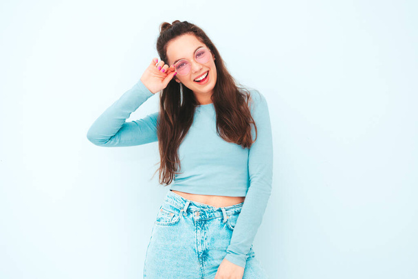 Young beautiful smiling female in trendy summer hipster clothes. Sexy carefree woman posing near light blue wall in studio. Positive model having fun indoors. Cheerful and happy - Photo, Image