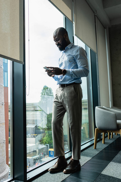 sorridente Africano americano empresário mensagens no smartphone perto da janela - Foto, Imagem