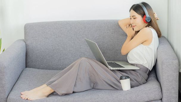 ノートパソコンで音楽を聴いている映画を見ているヘッドフォンを身に着けている女の子コーヒーを飲みながら休暇で自宅でソファの上で電話 - 写真・画像