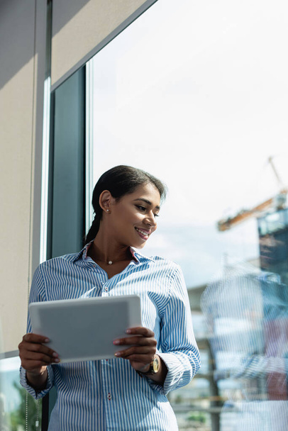 happy african american businesswoman looking through window while holding digital tablet - Φωτογραφία, εικόνα