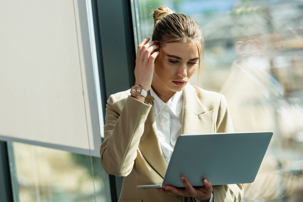 thoughtful businesswoman fixing hair while looking at laptop in office - Photo, Image