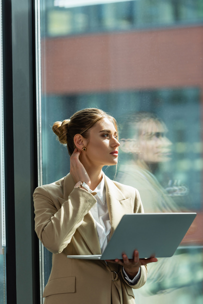 young businesswoman with laptop thinking near window in office - Foto, Imagem
