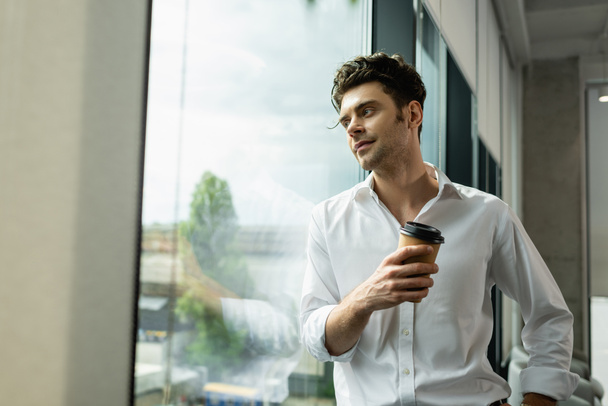 positive, dreamy businessman standing near window with coffee to go - Photo, Image