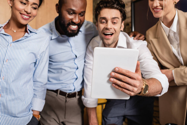 verbaasd zakenman houden digitale tablet in de buurt vrolijke interraciale collega 's - Foto, afbeelding