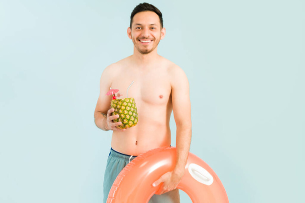 Handsome young man smiling and making eye contact while drinking a pina colada cocktail. Hispanic man in a swimsuit ready to go to the pool  - Photo, Image
