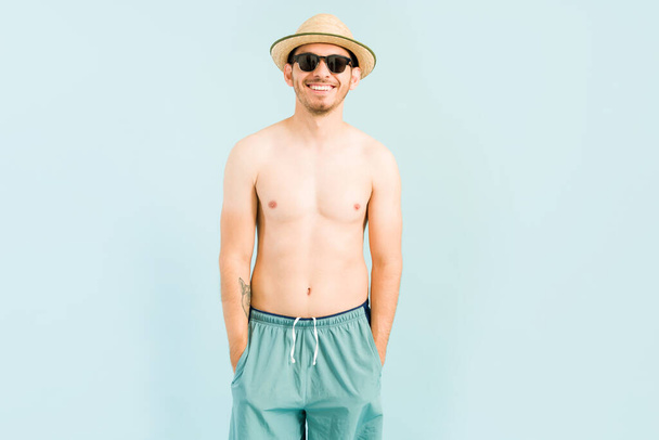 Είμαι έτοιμος να πάω να απολαύσω την παραλία. Όμορφος ευτυχισμένος άντρας με μαγιό, γυαλιά ηλίου και καλοκαιρινό καπέλο. Ο νεαρός χαμογελάει και ετοιμάζεται να πάει στην πισίνα. - Φωτογραφία, εικόνα
