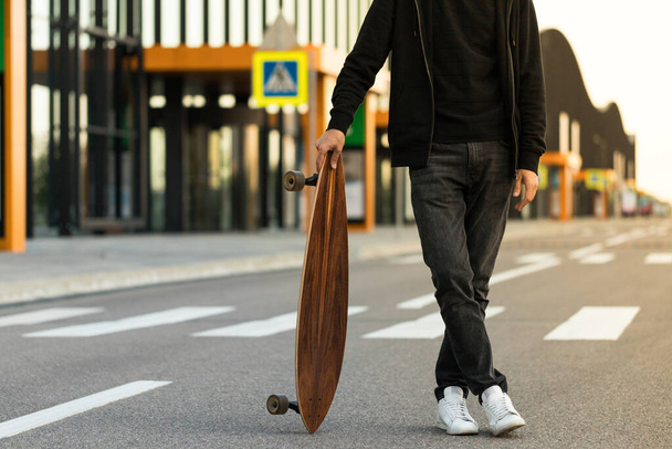 Человек хипстер, стоящий со скейтбордом или длинным бортом на открытом воздухе, обрезанное изображение. Досуг, здоровый образ жизни, экстремальные виды спорта - Фото, изображение
