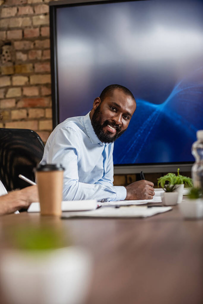 επιτυχημένος Αφροαμερικάνος επιχειρηματίας χαμογελώντας στην κάμερα κοντά στην οθόνη LCD στην αίθουσα συνεδριάσεων - Φωτογραφία, εικόνα