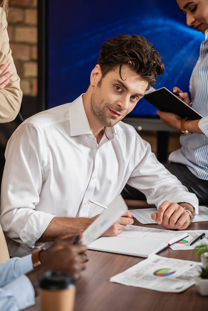 young businessman looking at camera while working with documents near blurred multiethnic coworkers - Photo, Image