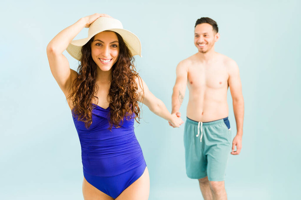 Retrato de una mujer atractiva sonriendo mientras se pone su sombrero de verano para evitar el sol en la playa. Novia cogida de la mano con su novio al lado de la piscina - Foto, imagen