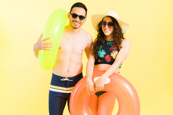 Divertida pareja joven en trajes de baño listos para ir a la playa. Novio y novia llevando flotadores y saliendo a la piscina en un día de verano - Foto, Imagen